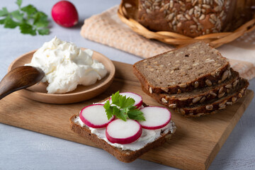 Fototapeta na wymiar Home made rye bread on a wooden cutting board with curd cheese, radish and ricotta and herbs. Decorated with green herbs