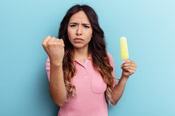 Young latin woman holding ice cream isolated on blue background showing fist to camera, aggressive facial expression.