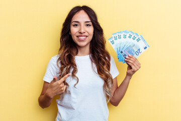 Young mixed race woman holding bills isolated on yellow background person pointing by hand to a shirt copy space, proud and confident