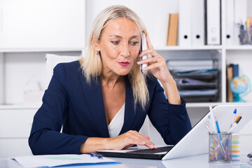 Smiling mature woman talking on mobile phone and using laptop in office