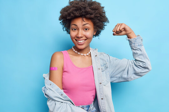 Photo of satisfied curly haired woman raises arm shows biceps wears pink t shirt denim shirt necklace adhesive bandage on shoulder after vaccination feels good and protected encourages to vaccinate