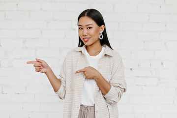 Brown-eyes woman in stylish cardigan abs beige pants smiles, looks into camera and points at place for text on white brick wall background.