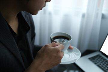 Closed up and cropped view with Asian businessman hold a cup of coffee and look on laptop computer.