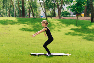 slim girl in a black gymnastic uniform performs gymnastic exercises or pilates on the green grass...