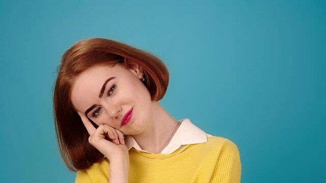 Young woman with red hair in yellow pullover looks straight propping chin with hand on blue background slow motion close view