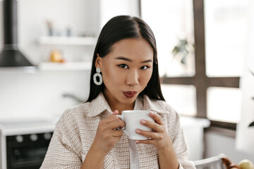 Close-up portrait of tanned brunette Asian woman with dark lipstick drinks tea. Lady looks into camera and holds white coffee cup in kitchen.