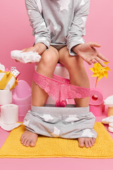 Faceless woman in pajama with slender legs holds napkin or diaper has lace panties pulled down on legs keeps bare feet on carpet isolated over pink background in restroom. Human necessities concept