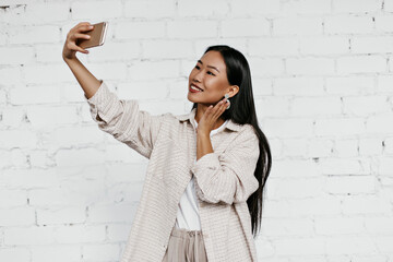 Cheerful brunette woman in beige cardigan smiles sincerely on white brick wall background. Tanned lady in good mood takes selfie.