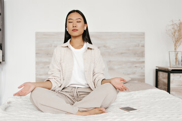 Brunette Asian woman in headphones meditating in bedroom. Charming lady in beige pants and cardigan sits on soft bed.