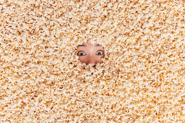 Surprised woman fully covered shows only eyes buriedin puffy popcorn cannot believe in shocking news. Cinema movies and entertainment. Female model buried in air popped corn spends free time at home