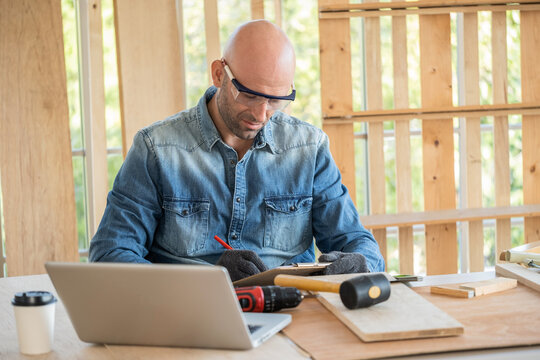 Professional carpenter man wearing glove and goggle writing some idea into clipboard in the modern wood workplace. Many equipment (hammer, screwdriver) on the table. Small business. DIY concept