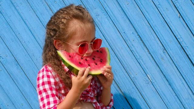 A beautiful cute girl in red sunglasses eats a sweet juicy piece of watermelon and licks her lips. National Watermelon Day. Eat and enjoy the flavors by smacking your lips