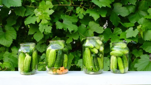 A woman's hand puts cucumbers in a jar for preserving vegetables in a marinade. There are  cans of cucumbers and carrots on the table. Pickled canned vegetables