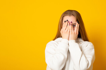 Banner. Scared young woman peeks through fingers, hides face, stares with opened eyes at something spooky, stands alarmed against yellow background, being troubled.