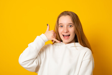 Positive young woman makes call me gesture keeps hands shaped like phone poses against yellow background. Cheerful girl asks for your telephone number.