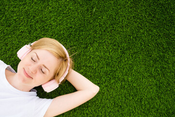 A young Caucasian girl in a white T-shirt and headphones lies on an artificial lawn with closed...