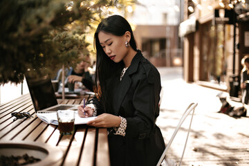 Charming young brunette lady in black trench coat make notes in notebook and works in laptop. Asian woman sits at wooden table outside.