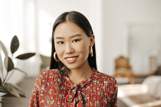 Close-up portrait of Asian brunette charming woman in red floral dress and massive earrings smiles and looks into camera in cozy room.