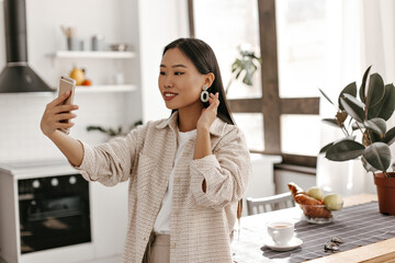 Cheerful tanned brunette woman in beige jacket takes selfie on kitchen. Asian lady in stylish outfit holds phone and smiles.