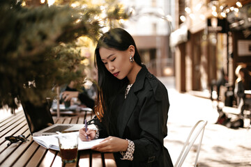 Pretty young lady in black trench coat write down information into notebook. Asian brunette woman works in laptop outside.