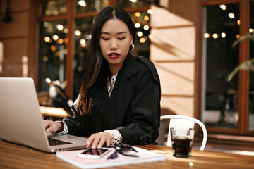 Portrait of brunette Asian woman in black jacket poses with laptop and coffee glass outside. Lady works outside and touches phone screen.