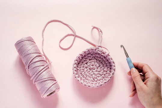 A hand holding a crochet hook and pink color yarn on a pink background, knitting and crochet supplies, hobby and craft