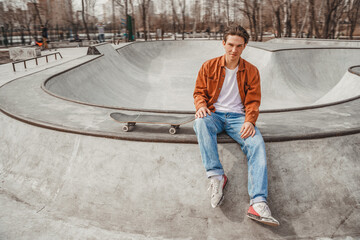 Skater boy sits on the edge of a ramp in a gray skatepark near a skateboard and looks straight...