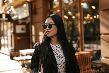 Obraz premium Brunette tanned Asian woman in stylish sunglasses, black trench coat and white blouse smiles. Portrait of attractive lady outside.