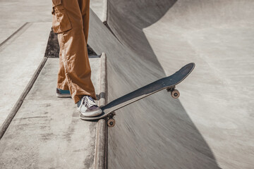 Skater in brown pants prepares for an important jump from a gray ramp in a skatepark, holding skates on the edge of the ramp, standing on it with his right foot