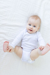 baby girl six months old plays with her feet lying on her back on a white bed in the bedroom of the house