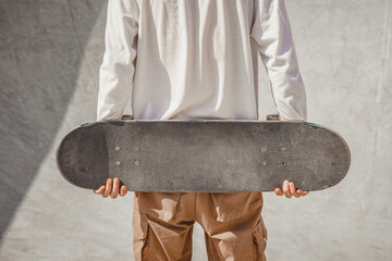 Skater boy in a white T-shirt and brown pants holding a skateboard behind his back preparing to jump on the ramp in a skatepark © Marharyta Hanhalo