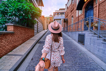 Follow me concept and traveling together. Girl traveler wearing hat and short jumpsuit holds the boyfriend's hand and walking around the european city - obrazy, fototapety, plakaty