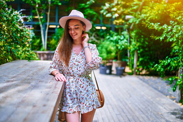 Portrait of young happy cheerful smiling cute beautiful brunette young girl with long hair wearing hat and short jumpsuit at green cafe at sunny summer day