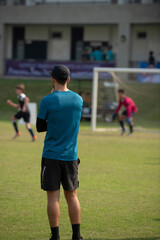Male youth football coach standing and watching his team playing in a school tournament on a clear sky and sunny day. Sport, active lifestyle, happy activity and coaching concept.	
