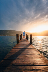 Orta San Giulio / Italy - June 2021: Young man at the bottom of a wooden jetty on Lake Orta as he watches the sun set behind the mountains at sunset