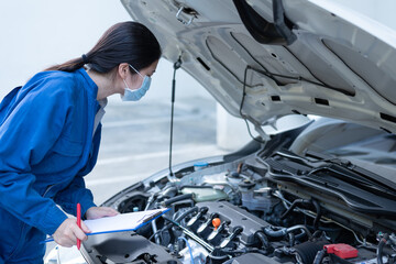 Medium shot of Asian woman holding clipboard, looking at engine, wearing blue jumpsuit and a mask, working, servicing, evaluating car condition. Female mechanic and car service maintenance concept.