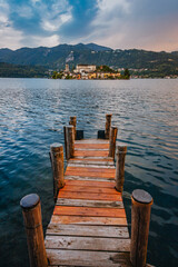 Naklejka premium Orta San Giulio / Italy - June 2021: The island of San Giulio with a wooden jetty in the foreground at sunset