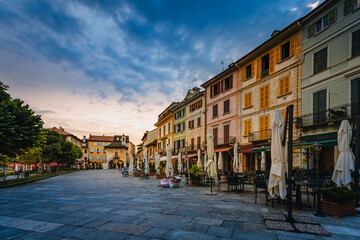 Fototapeta na wymiar Orta San Giulio / Italy - June 2021: Main square of the village of Orta San Giulio at sunset with blue clouds, with people sitting in the bars / restaurants of the square