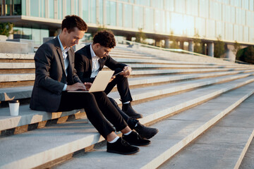 Businessmen young people work near the office in business suits holding gadgets in their hands. Successful colleagues work together. . They are discussing a new startup project.