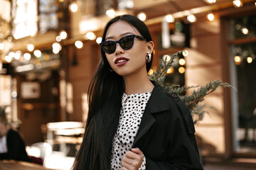 Cute tanned Asian woman in black trench coat and white dress poses outside. Portrait of brunette lady in sunglasses.