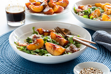 Close up of a bowl of grilled and peach and arugula salad with sunflower seeds against a bright sunny window.