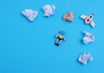 Top view of toy astronaut and crumpled paper like plates and clouds on a blue background.