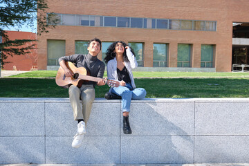 Young latin couple looking up to the sky and playing the guitar at the university campus. University life, millennial generation.