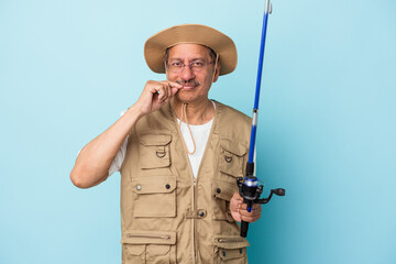 Senior indian fisherman holding rod isolated on blue background with fingers on lips keeping a secret.