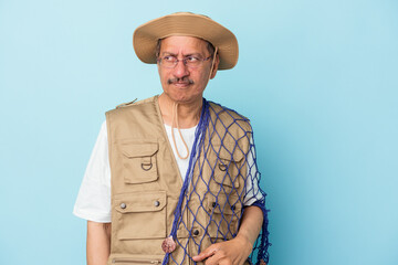 Senior indian fisherman holding net isolated on blue background confused, feels doubtful and unsure.