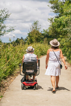 Rear view of an elderly woman on a disability scooter walking with her daughter on a path in a park