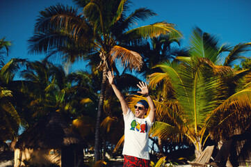 Happy attractive tourist man in white t shirt and sunglasses, walking with raised hands on the beach against palm trees background