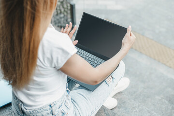 Laptop mockup. Young student girl uses laptop with white screen, sitting on stairs and looking at empty black monitor screen near office building. Copy space for design