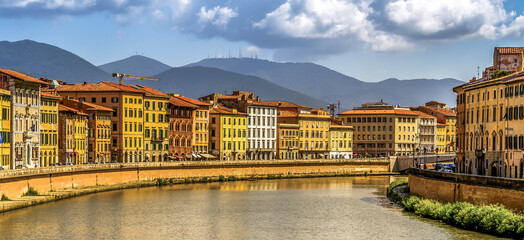 The Arno River flowing through Pisa, flanked by colorful buildings with the mountains in the background