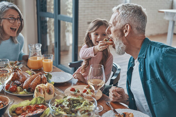 Happy little girl feeding her grandfather and smiling while having dinner with family